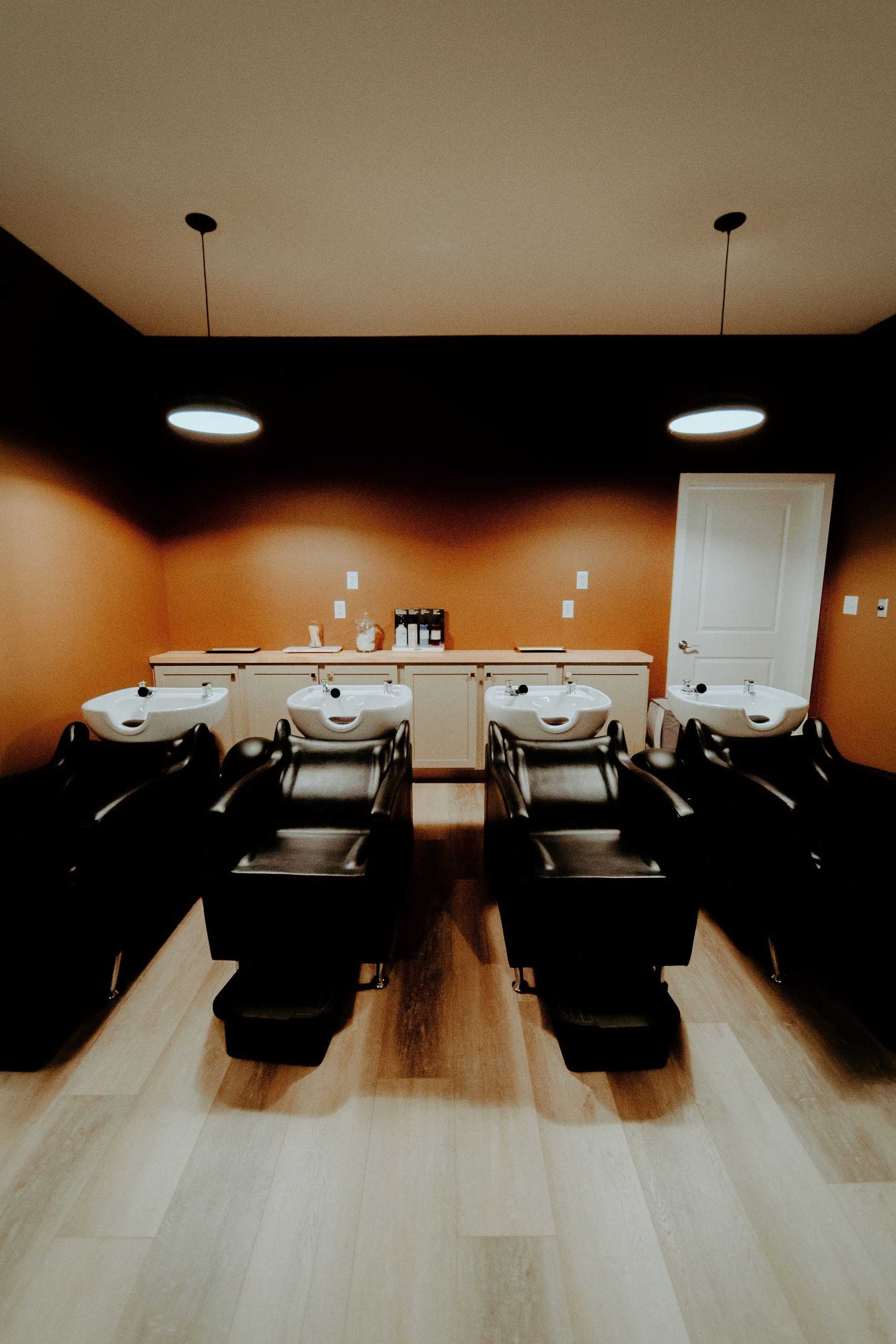 Modern hair salon with washing stations, black chairs, warm lighting, and wooden flooring.