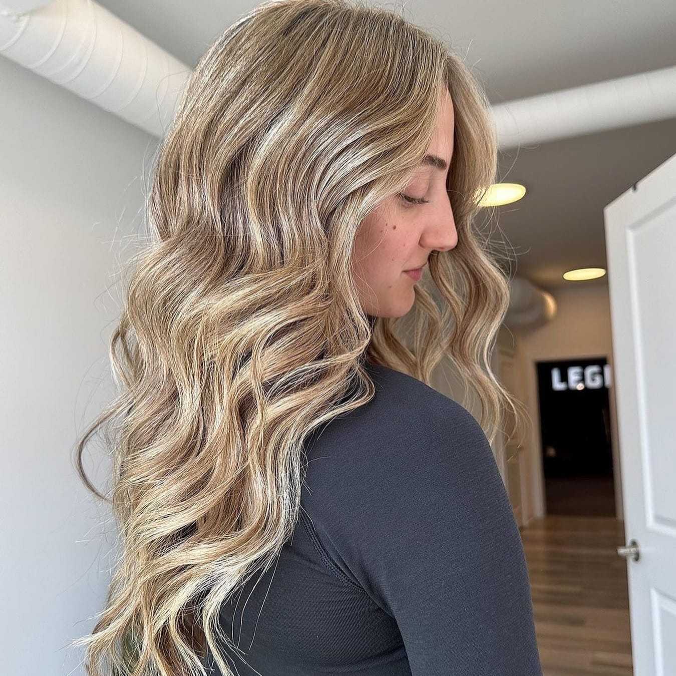 Woman with long, wavy blonde hair, wearing a black top, stands in a modern, well-lit room.